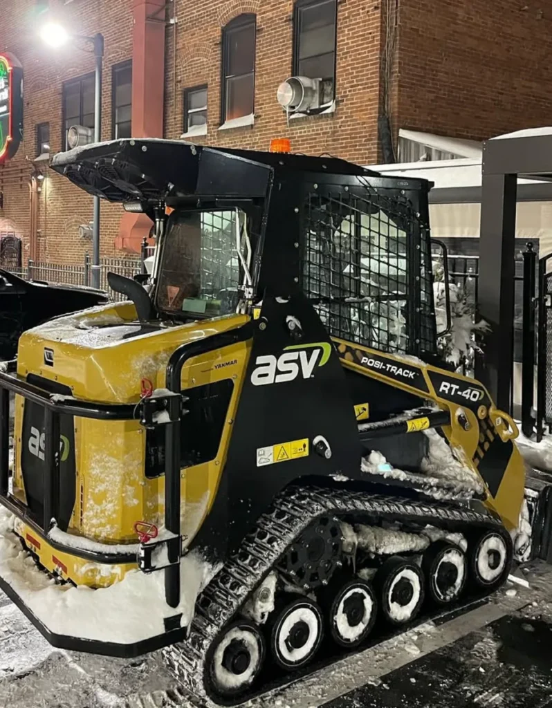 Compact tracked loader with snow on its tracks beside a brick building