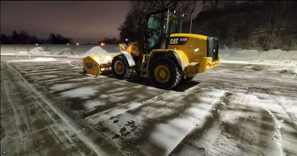 Wheel loader with a snow pusher on a snow-covered paved lot at night