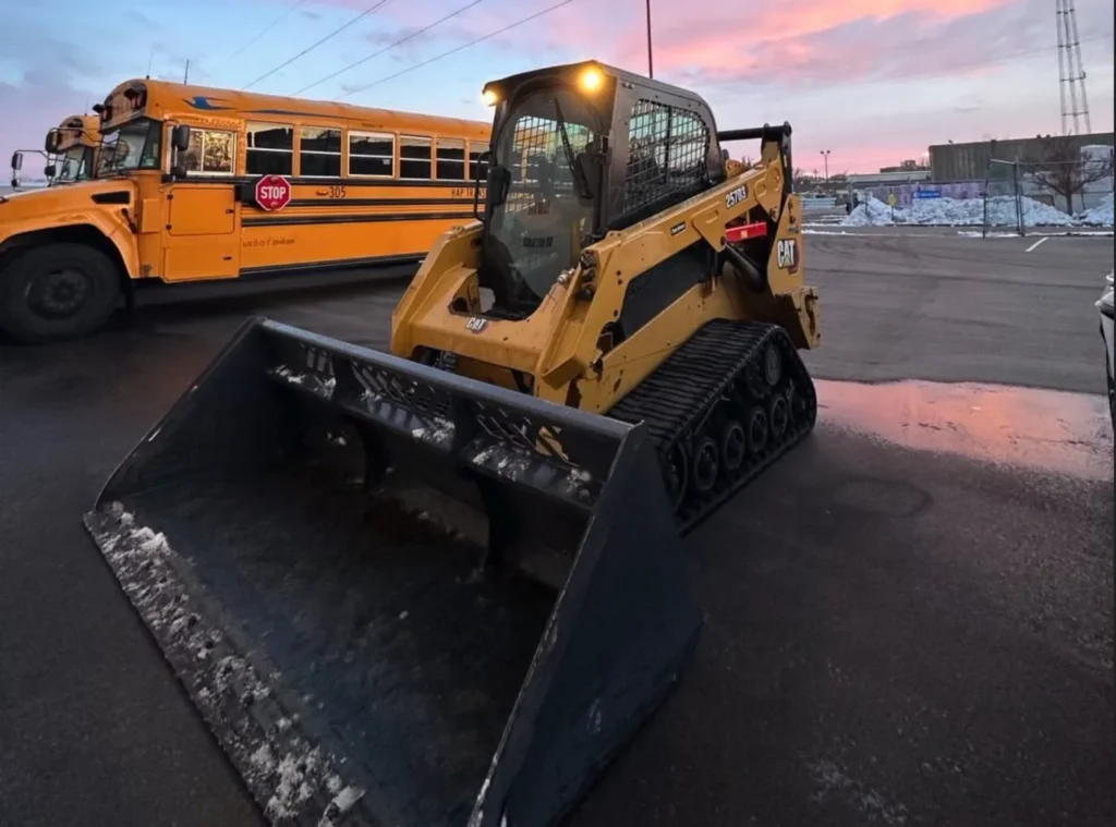 Tracked loader with a bucket on a cleared paved lot beside a school bus