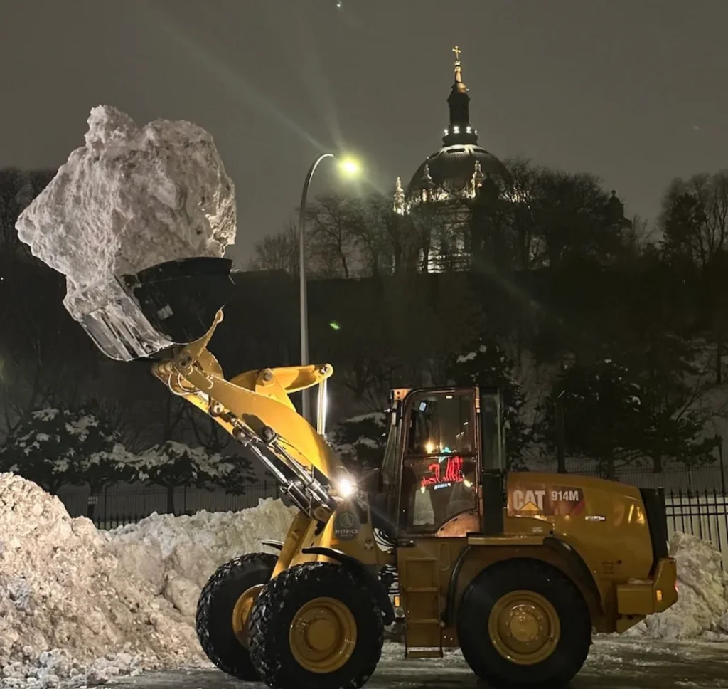 Wheel loader holding a raised bucket of snow beside a snowbank at night